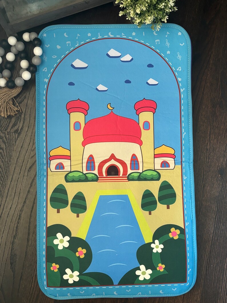 May include: A blue prayer mat featuring a cartoon mosque with red domes and yellow minarets. The design includes a blue sky with white clouds, a crescent moon, and musical notes. Green trees and flowers frame a blue river.