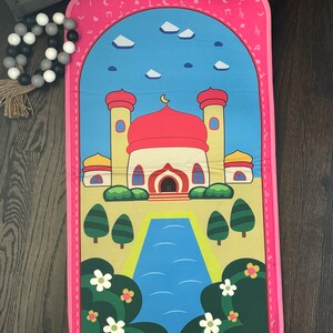 May include: A pink prayer mat featuring a cartoon mosque with a red dome, yellow walls, and a blue sky background. The mat has a pink border with musical notes and crescent moons.