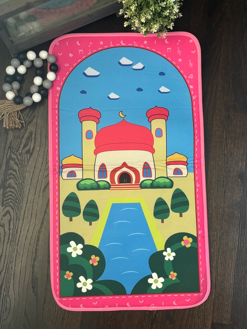 May include: A pink prayer mat featuring a cartoon mosque with a red dome, yellow walls, and a blue sky background. The mat has a pink border with musical notes and crescent moons.
