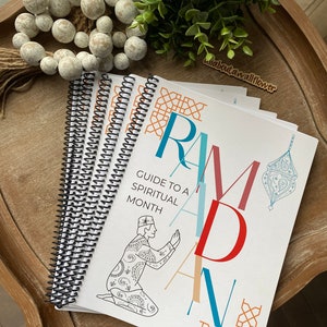 May include: A white and blue guide to a spiritual month with the word "Ramadan" in large colorful letters. The guide is spiral bound and features a black and white illustration of a person praying.
