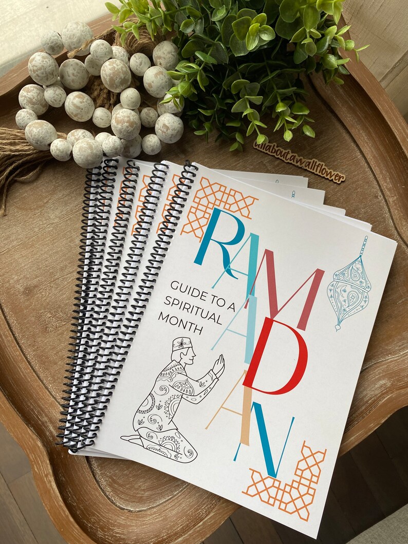 May include: A white and blue guide to a spiritual month with the word "Ramadan" in large colorful letters. The guide is spiral bound and features a black and white illustration of a person praying.