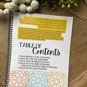 May include: A white and yellow spiral bound notebook with a table of contents for Ramadan prayers. The notebook has a gold background with Arabic text and a gold border.
