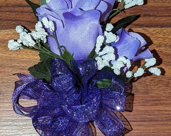 May include: A purple flower boutonniere with three purple roses and white baby's breath. The roses are surrounded by a purple sparkly bow.