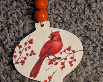 May include: Wooden Christmas ornament with a painted red cardinal bird perched on a branch with red berries. The ornament is topped with three red beads and a twine loop for hanging. The background is a textured gray surface.