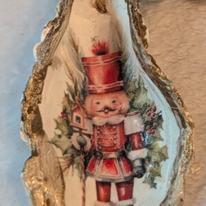 May include: An oyster shell Christmas ornament featuring a red and white nutcracker soldier design. The shell has a gold-painted edge and a jute string for hanging. Green and white beads are attached.