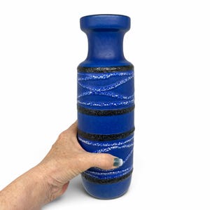 May include: A tall, cylindrical blue ceramic vase featuring a matte finish. The body is decorated with horizontal bands of textured black glaze and thin, wavy white speckled lines that wrap around the exterior of the vessel.