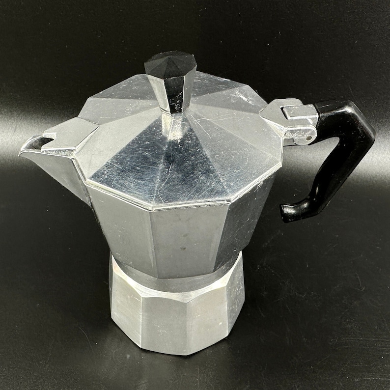 Puede incluir: Una cafetera de aluminio plateado con asa negra. La cafetera tiene una parte superior redondeada y una base hexagonal.