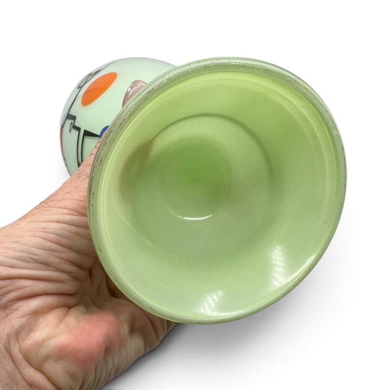 K&ouml;nnte beinhalten: Ein Eierbecher aus hellgr&uuml;nem Glas mit glatter, matter Oberfl&auml;che. Die Au&szlig;enseite zeigt ein grafisches Design mit schwarzen Linien, einem leuchtend orangefarbenen Kreis und einem blauen Punkt. Das Innere ist einfarbig hellminzgr&uuml;n mit einem breiten Rand.
