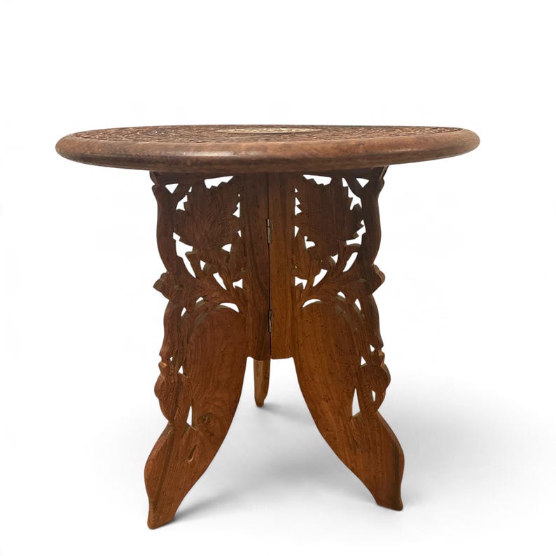 May include: A round wooden side table featuring a dark brown finish with intricate floral carvings on the tabletop and a folding base composed of three interlocking panels decorated with detailed leaf and vine relief patterns.