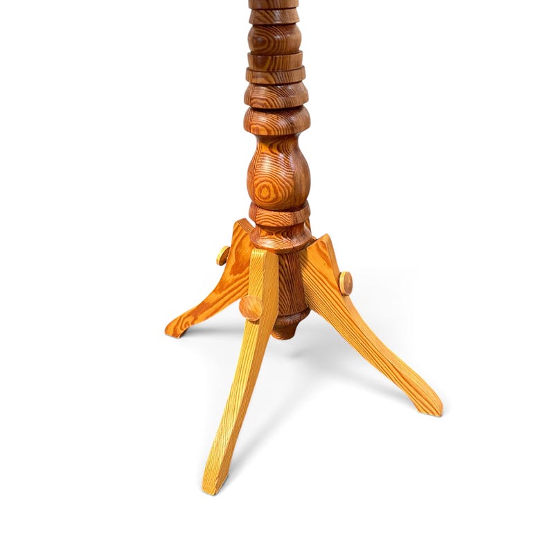 May include: A wooden stand featuring a tall, vertically stacked, turned-wood central column with a prominent grain pattern. The base consists of three angled, light-coloured wooden legs attached to the central post with circular fasteners.