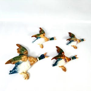 May include: A set of three ceramic mallard duck wall decorations in flight. Each duck features a glossy finish with painted details in shades of green, brown, white, and blue, depicting the birds with wings spread and beaks open in motion.