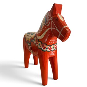Puede incluir: Una figura de caballo Dala de madera en color naranja brillante con patrones florales pintados a mano en blanco, verde y azul. El caballo muestra texto blanco en su costado que dice Avd.S Siljan, Avd 73 Fal, 50&aring;r y 1962, con un dise&ntilde;o de brida blanca.