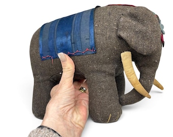 May include: A stuffed elephant toy made of brown textured fabric with two light tan tusks. A blue fabric panel is stitched onto the back with red thread. The elephant features decorative fabric accents on the head and ears, and yellow stitching on the feet.