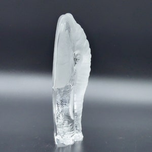May include: A clear, abstract glass sculpture with a frosted, textured surface. The sculpture has a curved, organic shape, with a smooth, frosted top and a textured, rougher base. The overall design is minimalist and modern.