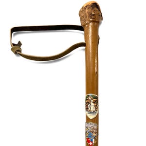 Puede incluir: Un bast&oacute;n de madera marr&oacute;n con un mango nudoso natural y una correa de cuero marr&oacute;n para la mu&ntilde;eca. El eje est&aacute; decorado con dos escudos met&aacute;licos de recuerdo, uno con la etiqueta Triberger Wasserf&auml;lle y el otro con Vierwaldst&auml;ttersee.
