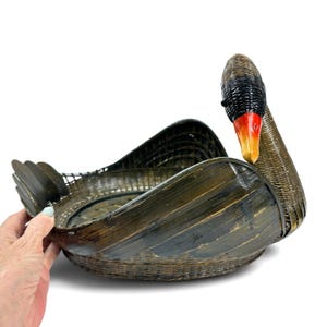 May include: A dark brown woven basket shaped like a swan with a long neck, a black head, and a bright orange beak. The body of the basket features a wide, open bowl area with curved wing-like sides and a small tail section at the back.