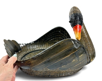 Puede incluir: Una cesta tejida de color marrón oscuro con forma de cisne, cuello largo, cabeza negra y pico naranja brillante. El cuerpo de la cesta presenta una zona de cuenco abierta y ancha con lados curvos en forma de ala y una pequeña cola en la parte posterior.