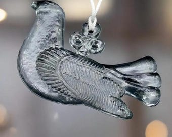 Peut inclure: Ornement en forme de colombe en verre transparent, avec une surface texturée et des détails sur les ailes et les plumes de la queue. La colombe est suspendue par une ficelle blanche et un charm décoratif en forme de trèfle. L'ornement mesure environ 10 cm de long.