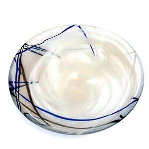 Pu&ograve; includere: Una ciotola in vetro rotonda e poco profonda con una base trasparente e motivi bianchi opachi che si intrecciano all'interno. Il bordo esterno &egrave; decorato con sottili linee incrociate in blu scuro e grigio scuro, creando un contrasto.