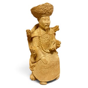 Op de afbeelding: Een beige uitgesneden beeldje van een zittende figuur met een versierde hoed en traditionele gewaden met complexe drakenmotieven. De figuur heeft een lange baard, houdt een klein voorwerp in de hand en zit op een stoel met uitgesneden armleuningen.