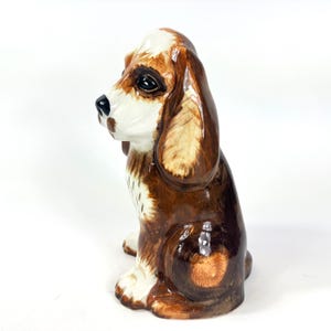 Peut inclure: Figurine en c&eacute;ramique repr&eacute;sentant un chien assis, avec des couleurs marron et blanc. Le chien a de longues oreilles tombantes et des traits faciaux d&eacute;taill&eacute;s. Objet d&eacute;coratif, probablement pour l'exposition.