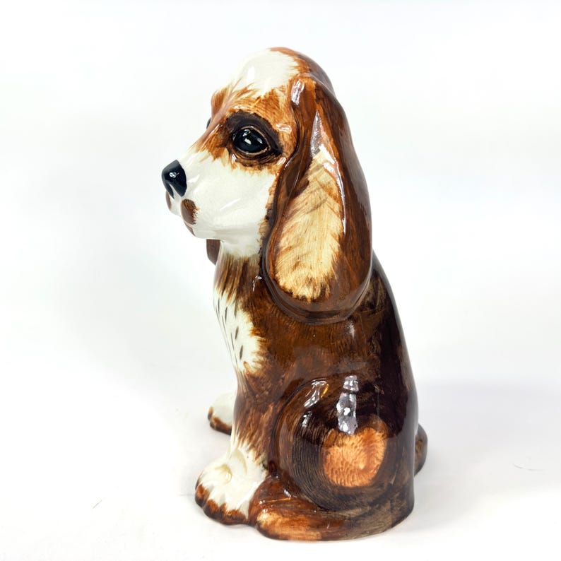Peut inclure: Figurine en c&eacute;ramique repr&eacute;sentant un chien assis, avec des couleurs marron et blanc. Le chien a de longues oreilles tombantes et des traits faciaux d&eacute;taill&eacute;s. Objet d&eacute;coratif, probablement pour l'exposition.