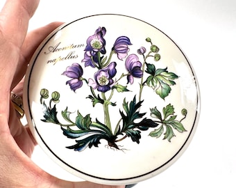 May include: A round ceramic lid featuring a botanical illustration of Aconitum napellus with purple flowers and green leaves on a cream background. The text Aconitum napellus is printed in black script on the left side of the floral design.