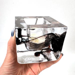 Könnte beinhalten: Ein transparenter, würfelförmiger Glaskerzenhalter mit dicken Wänden und einer eisartigen Textur. Das Glas enthält dunkle, abstrakte schwarze Einschlüsse und trägt den Schriftzug Kosta Boda auf der unteren Vorderseite des Objekts.