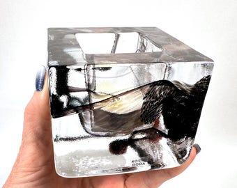 May include: A clear, cube-shaped glass candle holder with thick walls and a textured, ice-like appearance. The glass features dark, abstract black inclusions within the structure and the text Kosta Boda printed on the lower front surface.