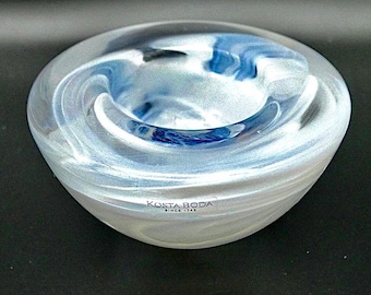 May include: A Kosta Boda glass bowl with a swirling design of white and blue. The bowl has a rounded shape with a central depression. The text "KOSTA BODA SINCE 1742" is visible on the side.