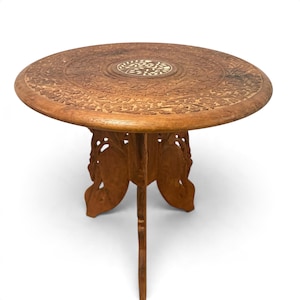 May include: A round wooden side table featuring intricate floral carvings across the tabletop surface. The center of the table displays a circular inlay with a light-colored leaf pattern. The base consists of three carved wooden legs.