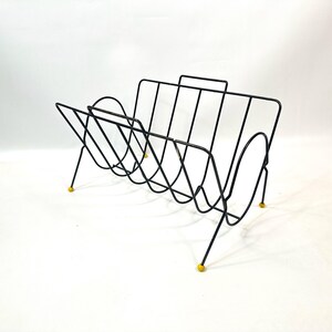 May include: A black wire magazine rack with a mid-century modern design. The rack has a handle and is supported by thin legs with yellow tips. The rack is designed to hold magazines and other reading materials.