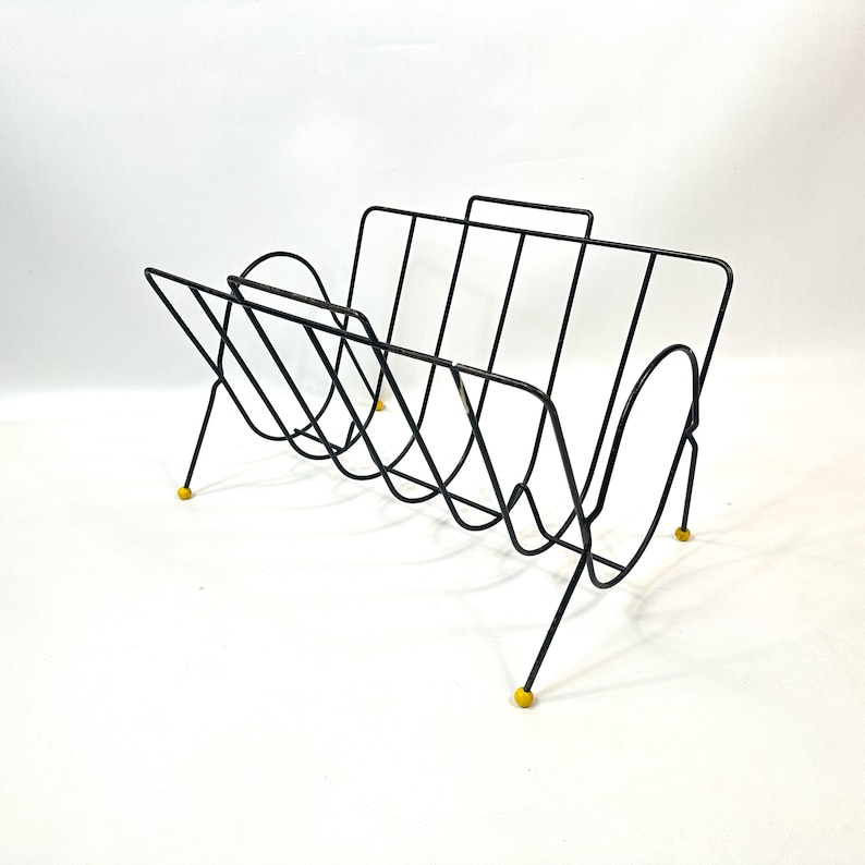 May include: A black wire magazine rack with a mid-century modern design. The rack has a handle and is supported by thin legs with yellow tips. The rack is designed to hold magazines and other reading materials.