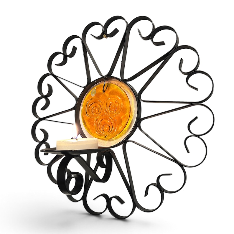 Peut inclure: Une applique murale en m&eacute;tal noir dot&eacute;e d'un cadre circulaire avec des volutes d&eacute;coratives et un disque en verre ambr&eacute; au centre. Un petit bougeoir circulaire est fix&eacute; &agrave; l'avant, contenant une bougie chauffe-plat blanche avec une flamme.