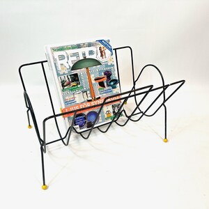 May include: Black metal magazine rack with a geometric design, holding a magazine with colorful images. The rack has yellow feet and a mid-century modern aesthetic.
