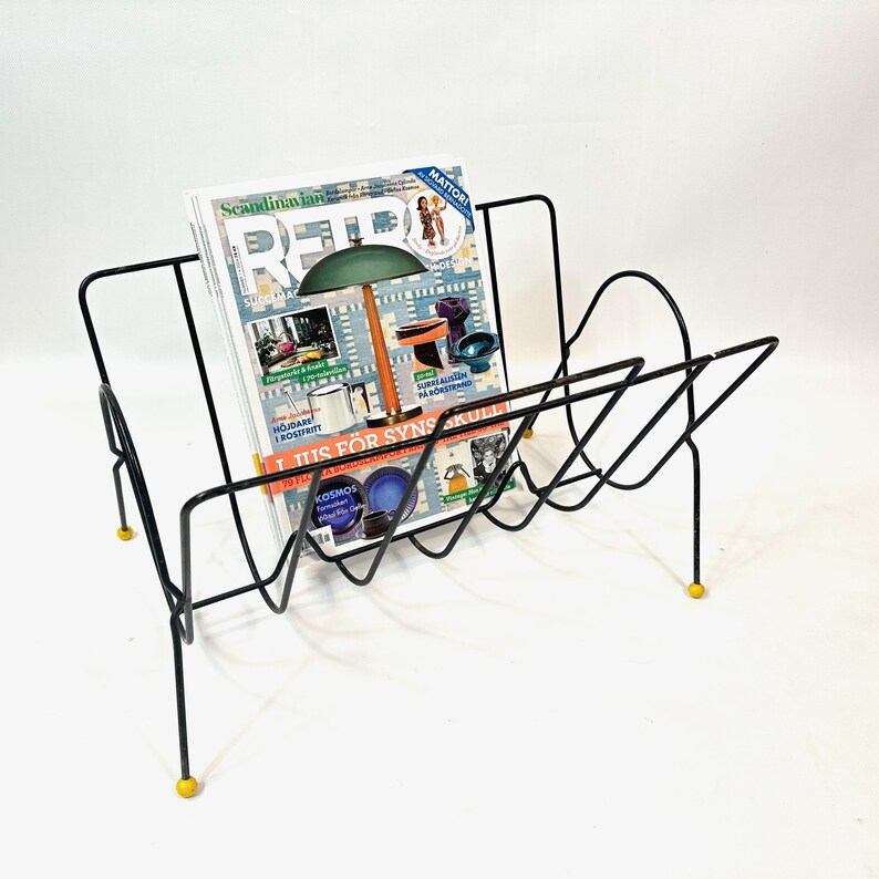 May include: Black metal magazine rack with a geometric design, holding a magazine with colorful images. The rack has yellow feet and a mid-century modern aesthetic.