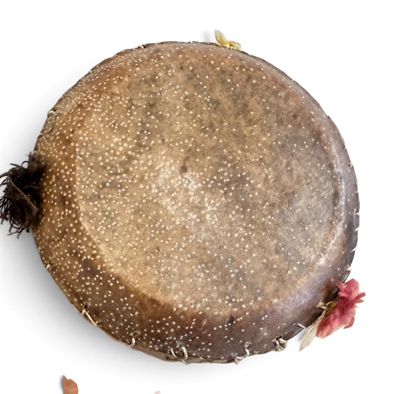 May include: A circular, brown, textured drum with a speckled white pattern across its surface. The edges are secured with light-coloured stitching, and it features a dark brown tassel on the left side and a small pink floral accent on the right side.