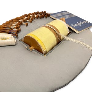 May include: A circular gray lace pillow featuring a central yellow cylindrical bolster, numerous wooden bobbins with white thread, a spool of white thread, and a blue book titled Knyppling. The setup is designed for bobbin lace making.