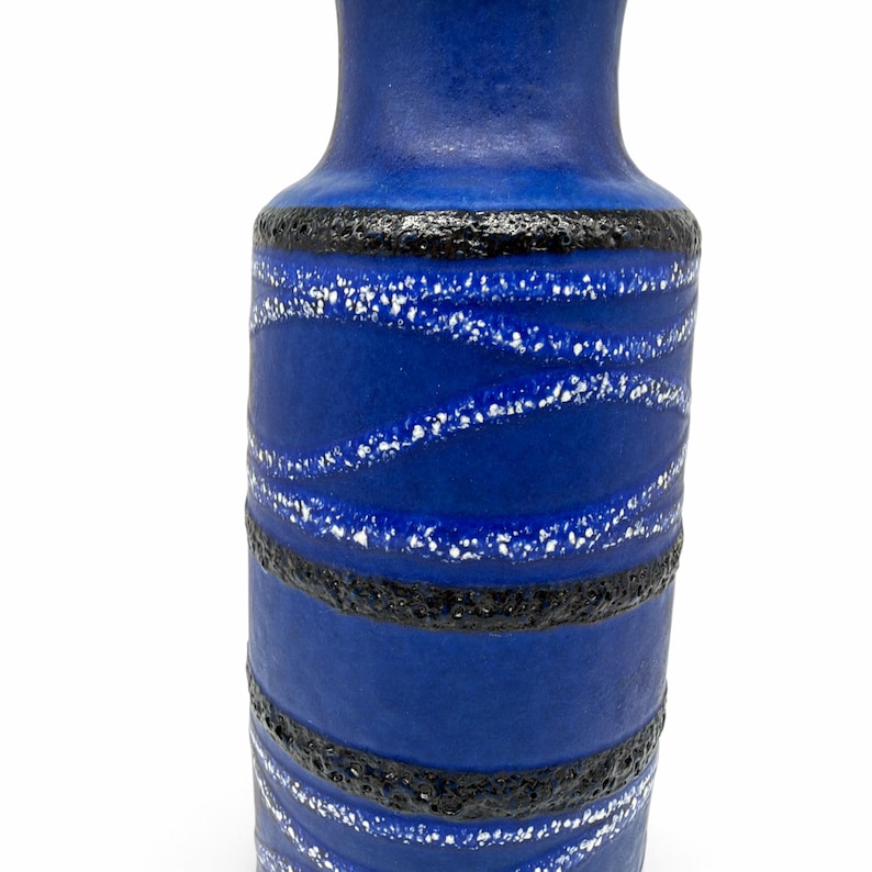 May include: A cylindrical ceramic vase featuring a vibrant blue matte finish. The surface is decorated with horizontal bands of textured black lava glaze and wavy, diagonal lines of white speckled glaze that wrap around the body.