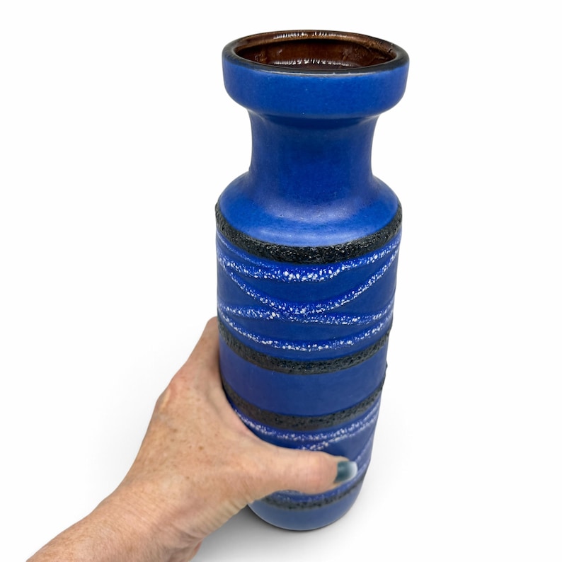 May include: A tall cylindrical ceramic vase with a vibrant blue matte finish. The exterior features horizontal bands of textured black glaze interspersed with thin, wavy white lines that wrap around the body. The interior rim is dark brown.