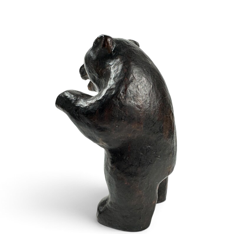 May include: A dark brown bear figurine standing on its hind legs with its front paws raised. The sculpture features a textured, uneven surface with visible brushstrokes or molding marks, capturing the bear in a dynamic, upright posture.