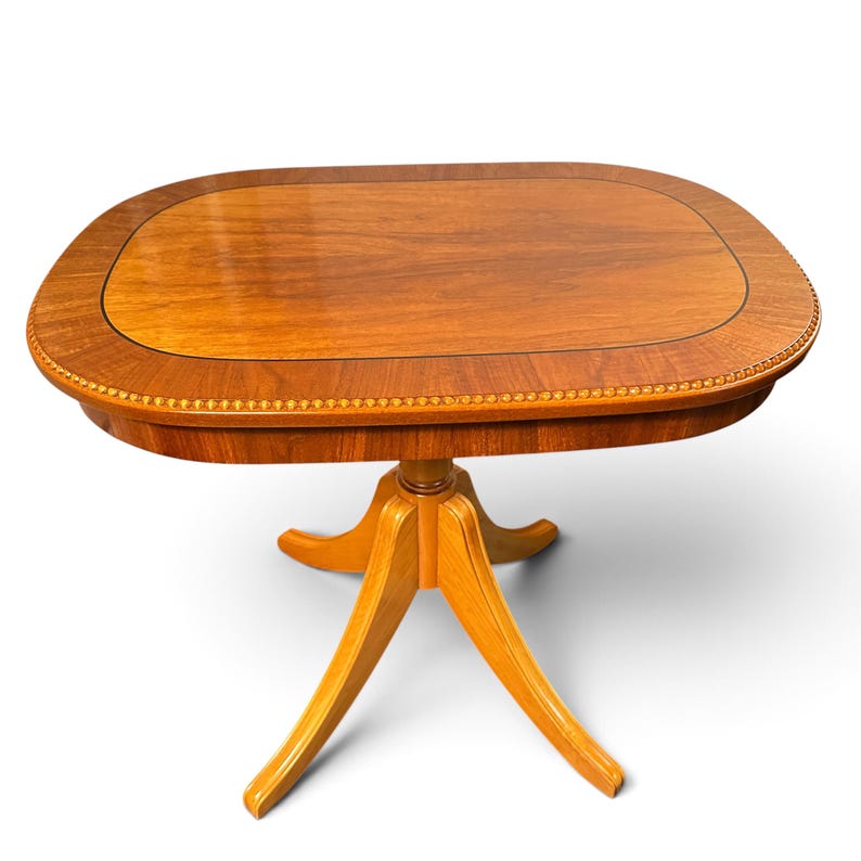 Op de afbeelding: Een houten zuiltafel met een afgerond rechthoekig blad met een glad, gepolijst bruin oppervlak en een decoratieve kralenrand. De tafel wordt ondersteund door een centrale kolom die uitloopt in vier gebogen poten aan de basis.
