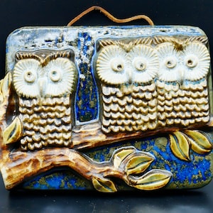 May include: A rectangular ceramic wall hanging featuring three owls perched on a branch. The owls have white and tan feathers, with large eyes. The background is blue and the branch is brown. A leather strap is attached for hanging.