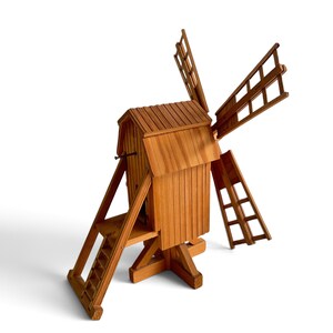 May include: A wooden model of a windmill featuring a vertical main structure with a gabled roof, a set of four lattice-style blades, and an attached staircase leading to the entrance. The entire structure is crafted from light brown wood.