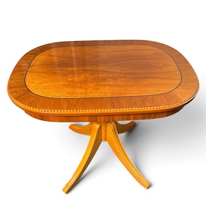 Op de afbeelding: Een houten zuiltafel met een glad, ovaalvormig blad met een gepolijste afwerking en een decoratieve kralenrand. De tafel wordt ondersteund door een centrale kolom die uitloopt in vier gebogen poten, alles in een warme, lichtbruine houttint.