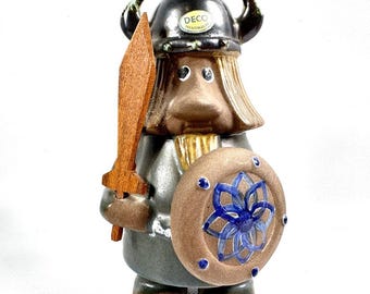 May include: A ceramic figurine of a warrior wearing a black horned helmet with a DECO HANDMADE sticker. The figure has brown hair, a brown beard, and a gray tunic. It holds a wooden sword in one hand and a round brown shield with a blue floral pattern.