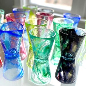 Puede incluir: Colecci&oacute;n de vasos de chupito soplados a mano, coloridos. Los vasos presentan una variedad de colores, incluyendo azul, verde, rosa y negro, con dise&ntilde;os en espiral. Se exhiben sobre una superficie reflectante, mostrando sus dise&ntilde;os &uacute;nicos.