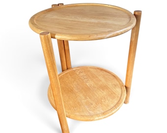 Könnte beinhalten: Ein hellbrauner Beistelltisch aus Holz mit zwei runden Ebenen und erhöhten Kanten. Der Tisch wird von drei zylindrischen Beinen gestützt, die vertikal verlaufen und die obere sowie die untere Ablage in einer einfachen Struktur verbinden.