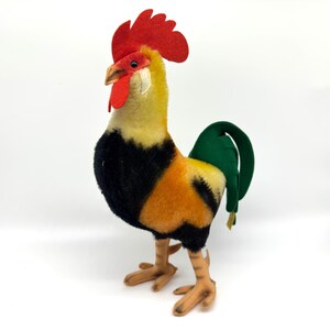 May include: A plush rooster figurine with a red comb and wattle, yellow and orange body, black chest, and green tail. The rooster has tan legs and feet, and a tan beak. The figurine is set against a white background.