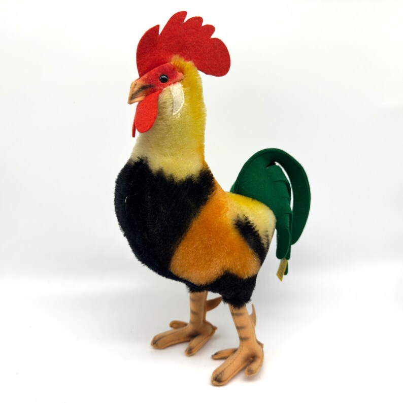 May include: A plush rooster figurine with a red comb and wattle, yellow and orange body, black chest, and green tail. The rooster has tan legs and feet, and a tan beak. The figurine is set against a white background.
